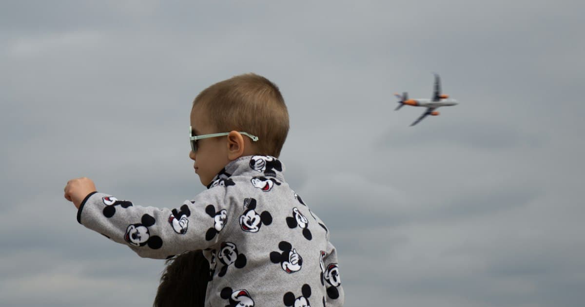Un petit garçon en lunettes de soleil, sur les épaules d'un adulte, montre du doigt un avion qui décolle